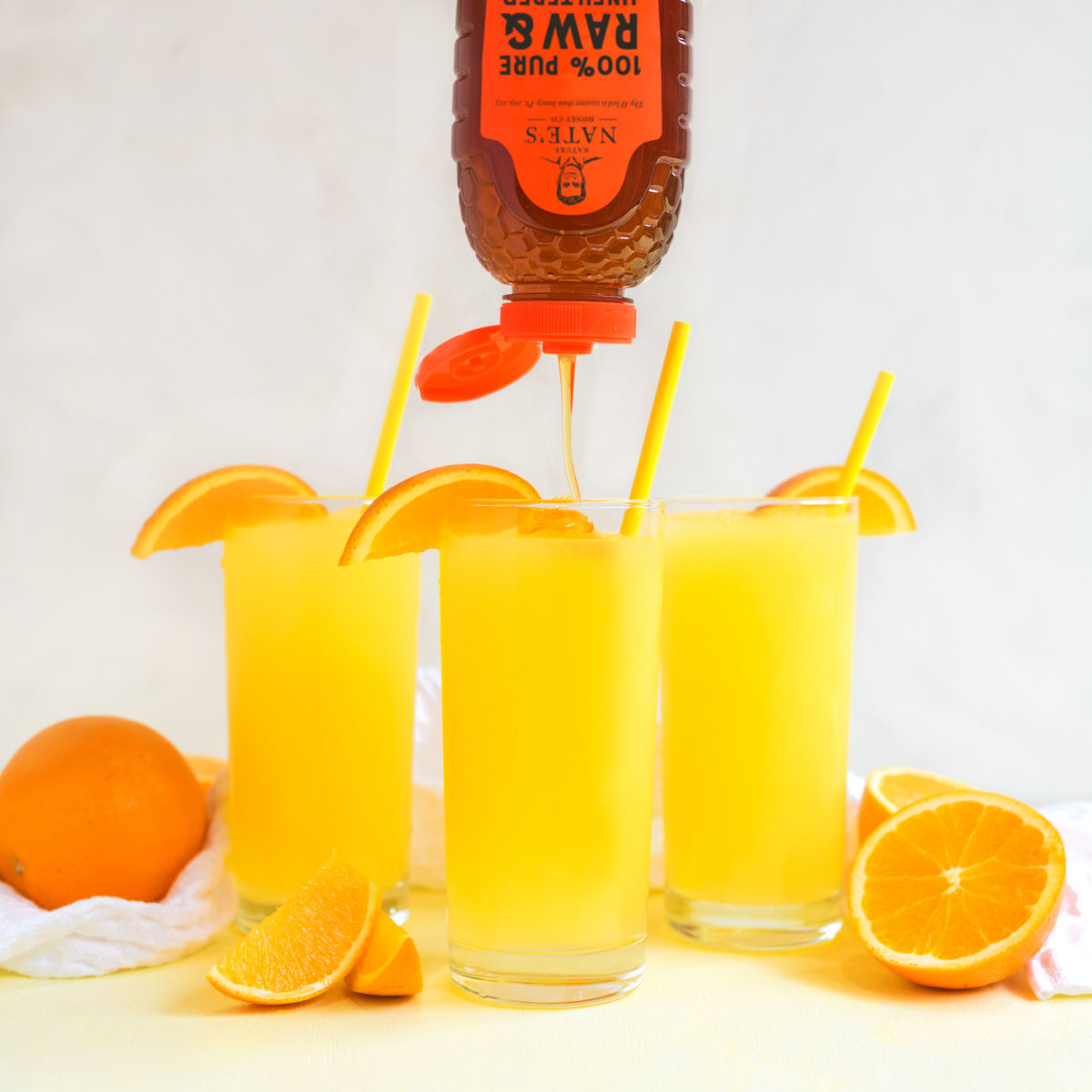 Three glasses of citrus slushie with straws and orange slices with Nate's honey being drizzled into one.
