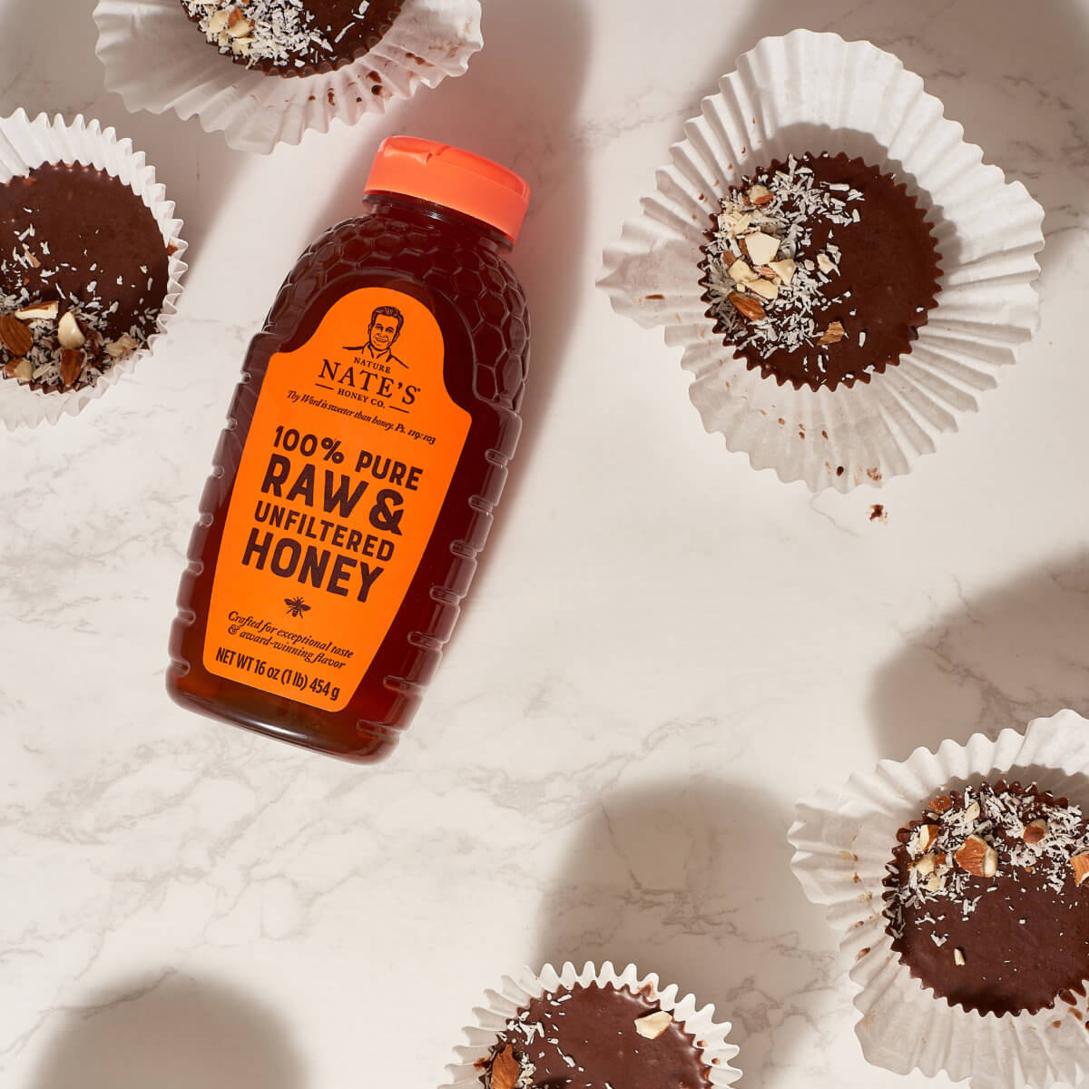 A bottle of Nate’s honey surrounded by chocolate coconut almond treats on a marble surface.