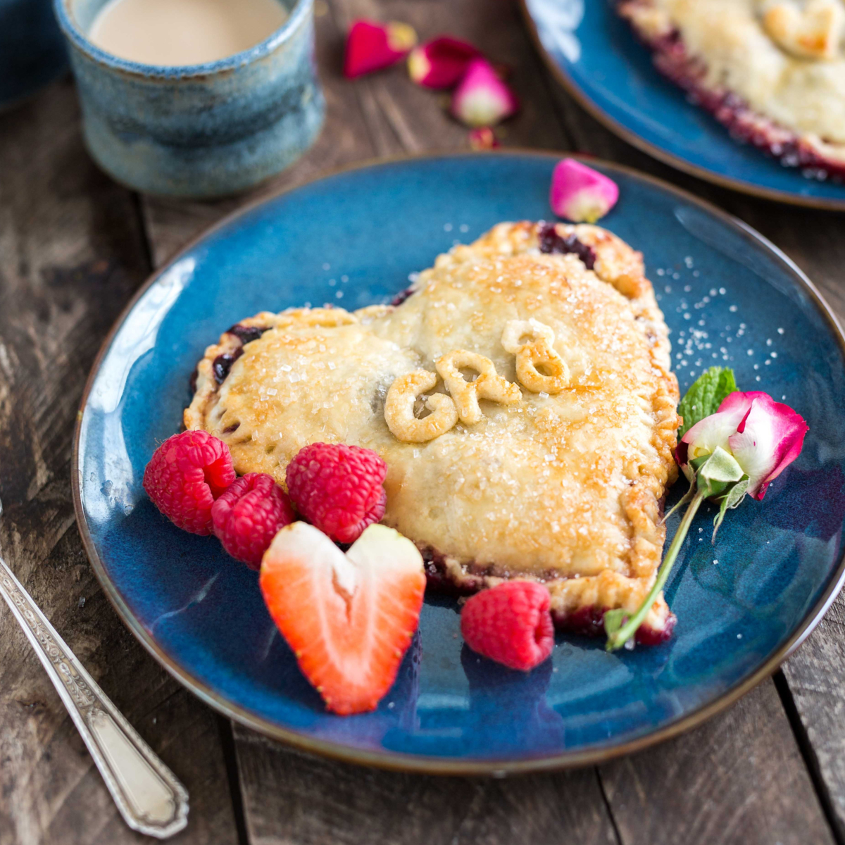 Heart-shaped tart on blue plate with berries and a side of coffee, rustic wooden table setting.