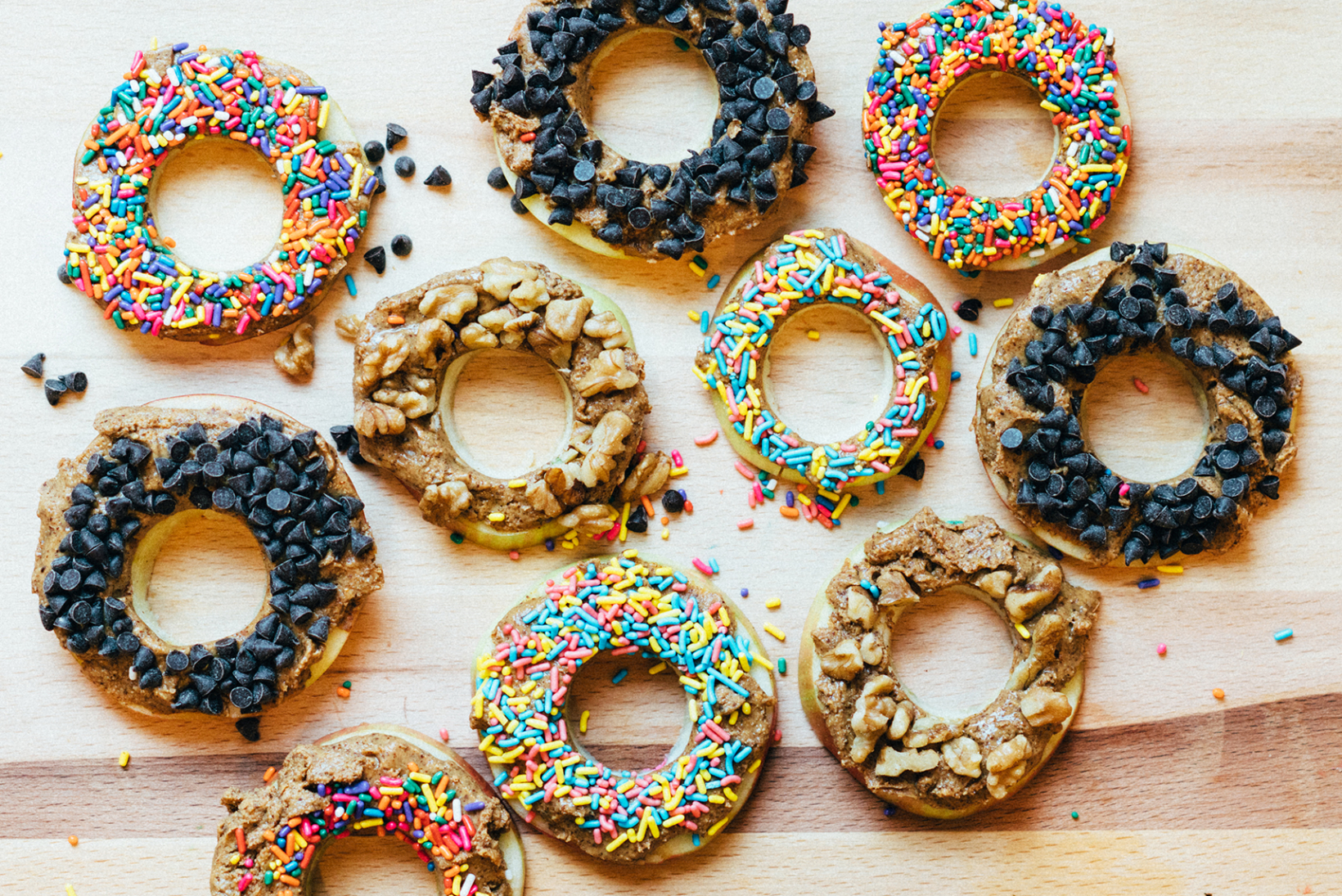 Apple rings topped with nut butter, chocolate chips, sprinkles and nuts on wood.