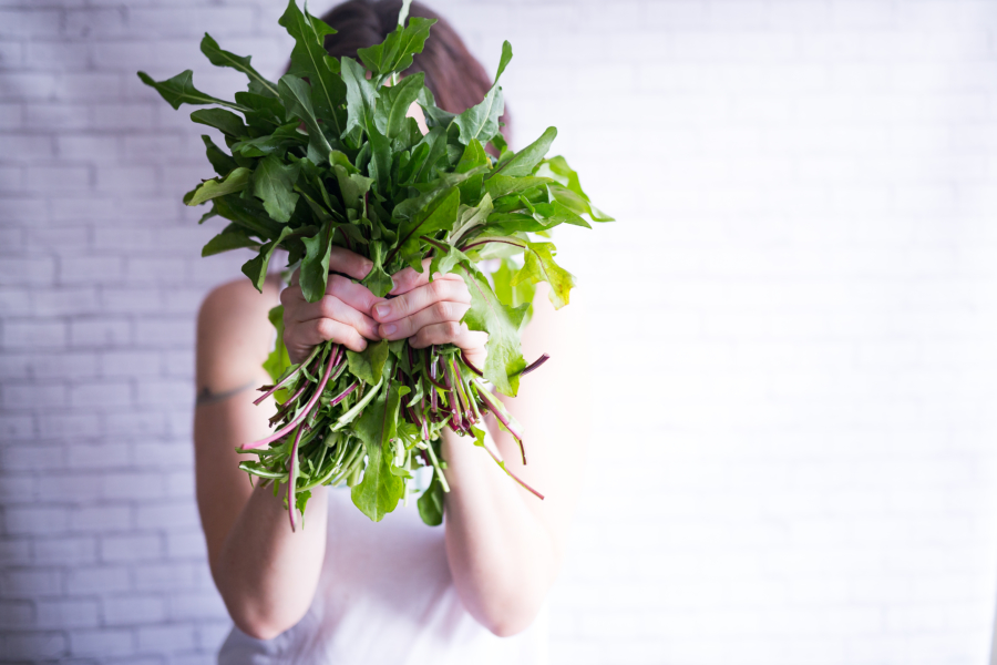 Person holding a large bunch of leafy greens in front of their face against a white brick wall.
