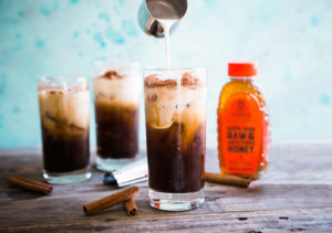 Pouring cream into iced coffee, with more glasses, cinnamon, and honey bottle.