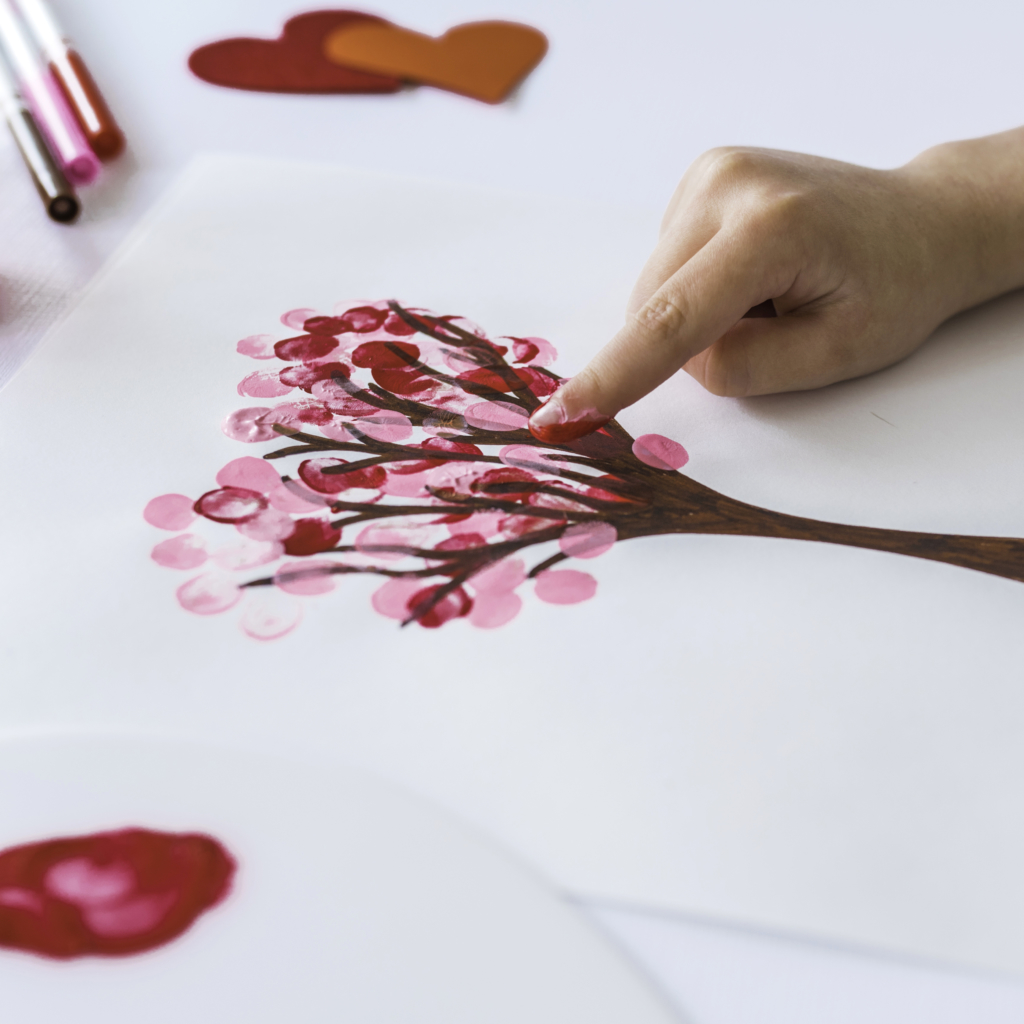 A hand painting a pink heart tree with fingerprint leaves, with art supplies and cut-out hearts around.