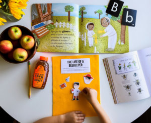 Child's hands with beekeeping book, honey bottle, apples, sunflowers on a yellow surface.