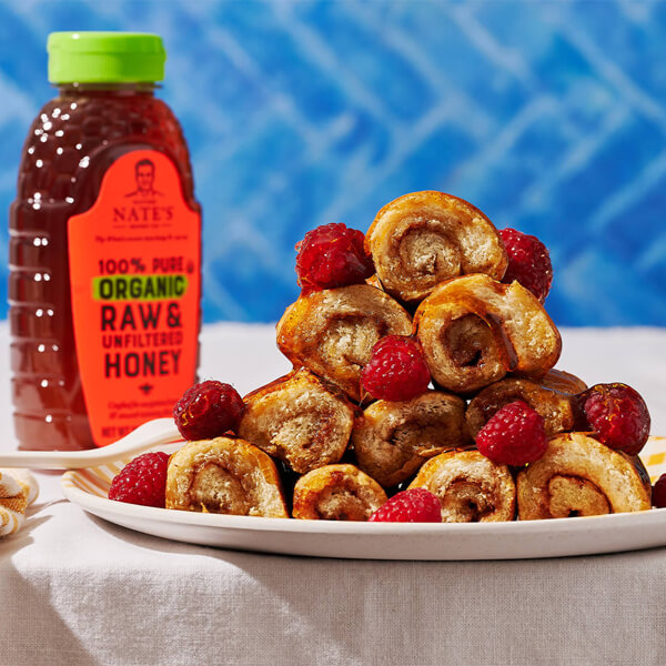 Plate of French Toast Rollups with fresh raspberries, served alongside a bottle of Nate's 100% Pure Organic Raw & Unfiltered Honey.