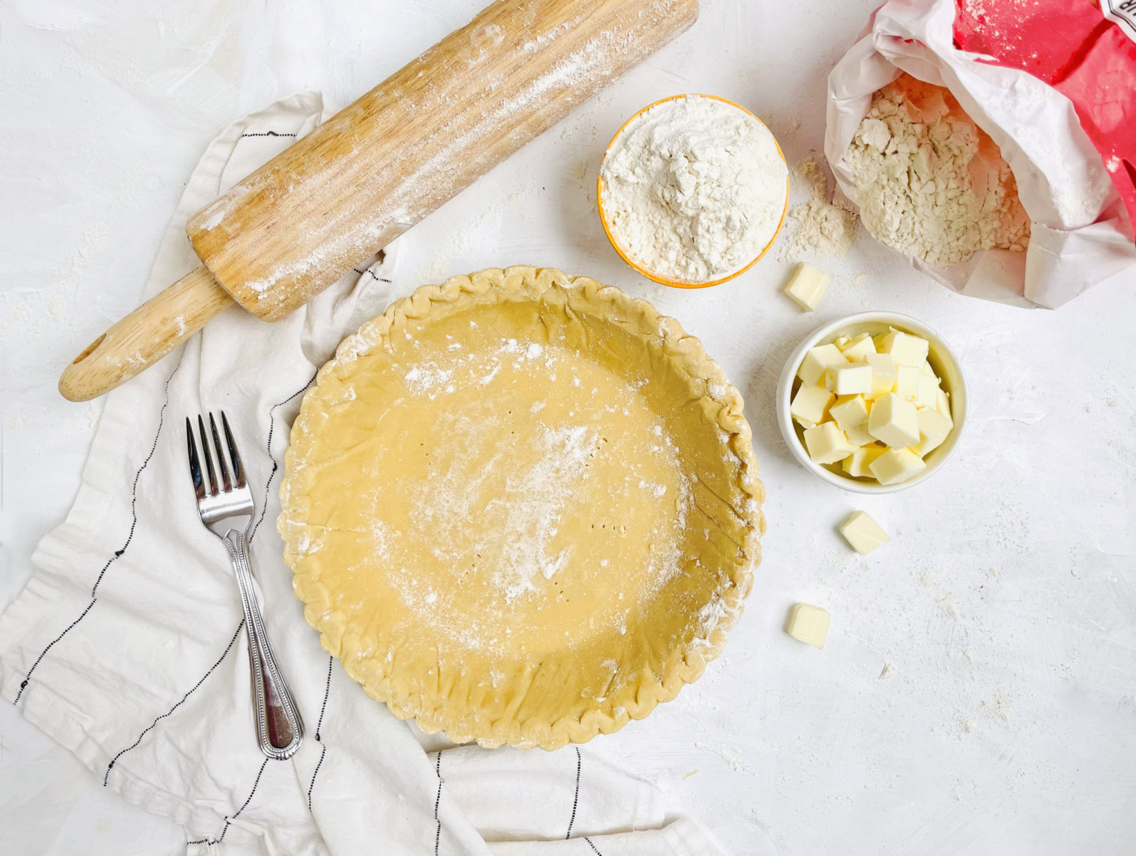 Unbaked pie crust on a white surface, with flour, butter cubes, a rolling pin, and a fork nearby.