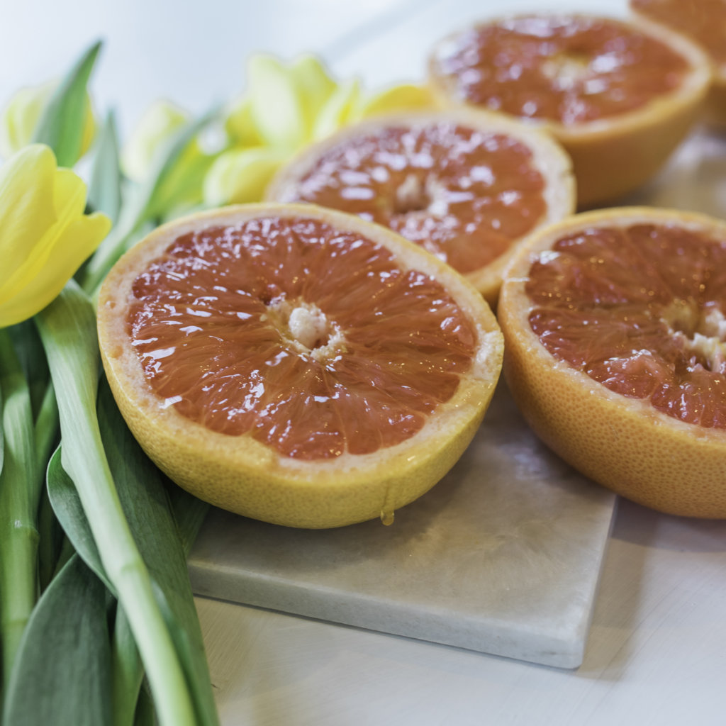 Sliced grapefruits drizzled with honey beside yellow tulips on a marble board.