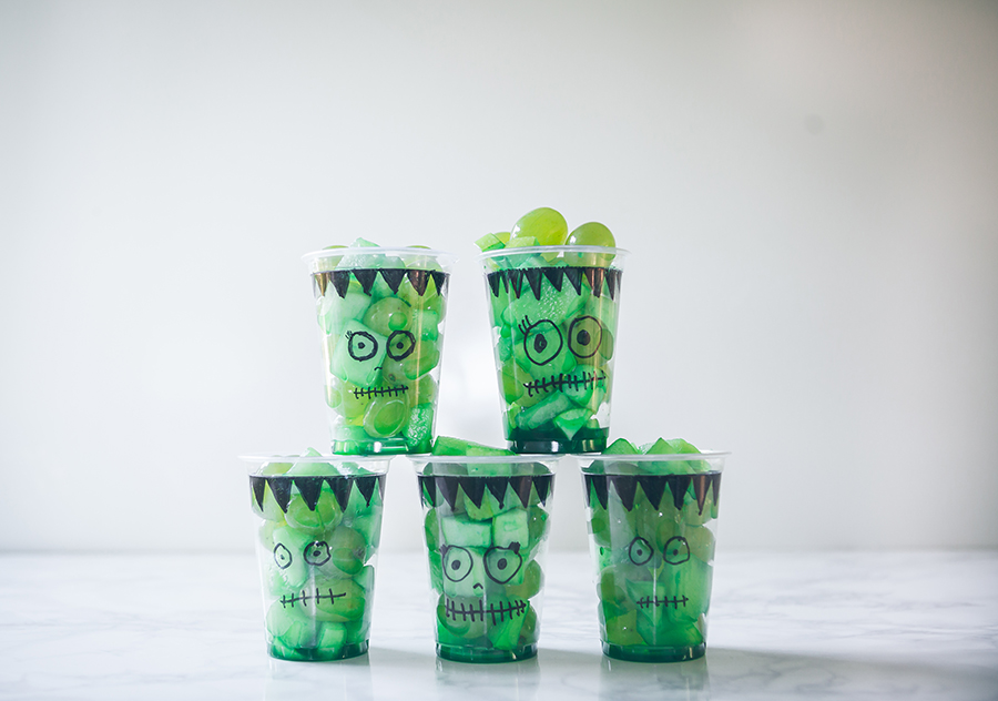 Frankenstein themed cups filled with green fruit pieces in a row on white marble, plain wall backdrop.