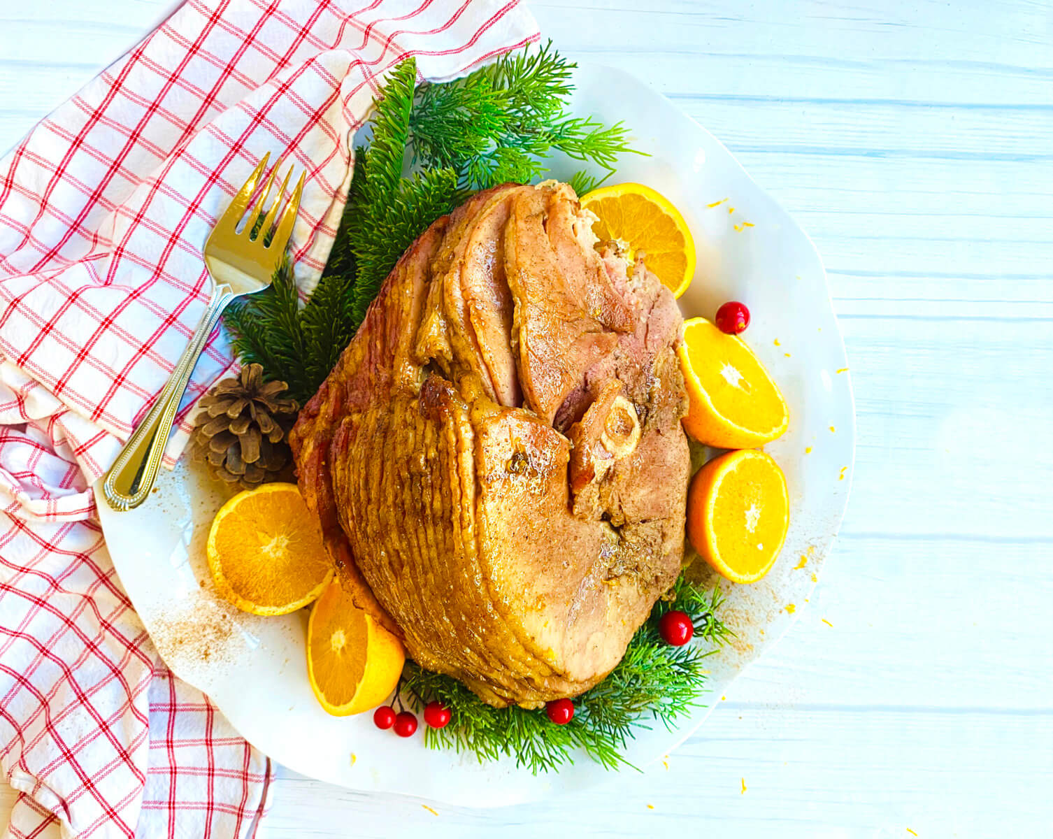 A baked ham on a white platter garnished with orange slices, green herbs, and red berries, with a red checkered napkin.