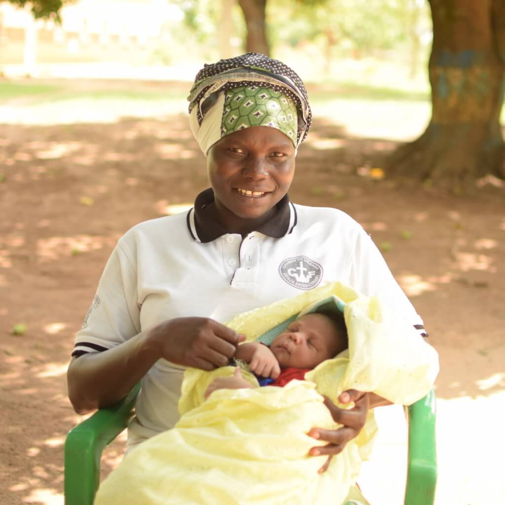A woman in a headscarf smiling, holding a sleeping baby wrapped in a yellow blanket, seated outdoors. A woman in a headscarf smiling, holding a sleeping baby wrapped in a yellow blanket, seated outdoors.