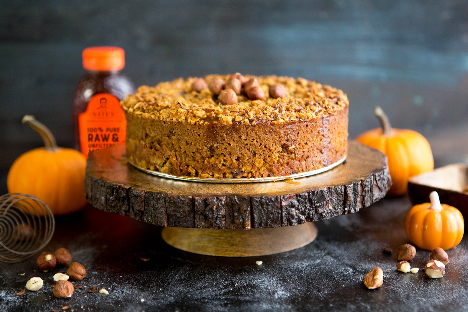 A hazelnut and pumpkin pie on a wood slice stand, with a Nate’s honey bottle and pumpkins, on a dark backdrop.