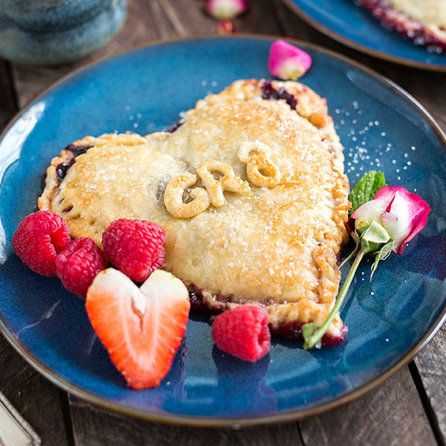 Heart-shaped berry hand pie on a blue plate with fresh strawberries and raspberries, on a wooden table.