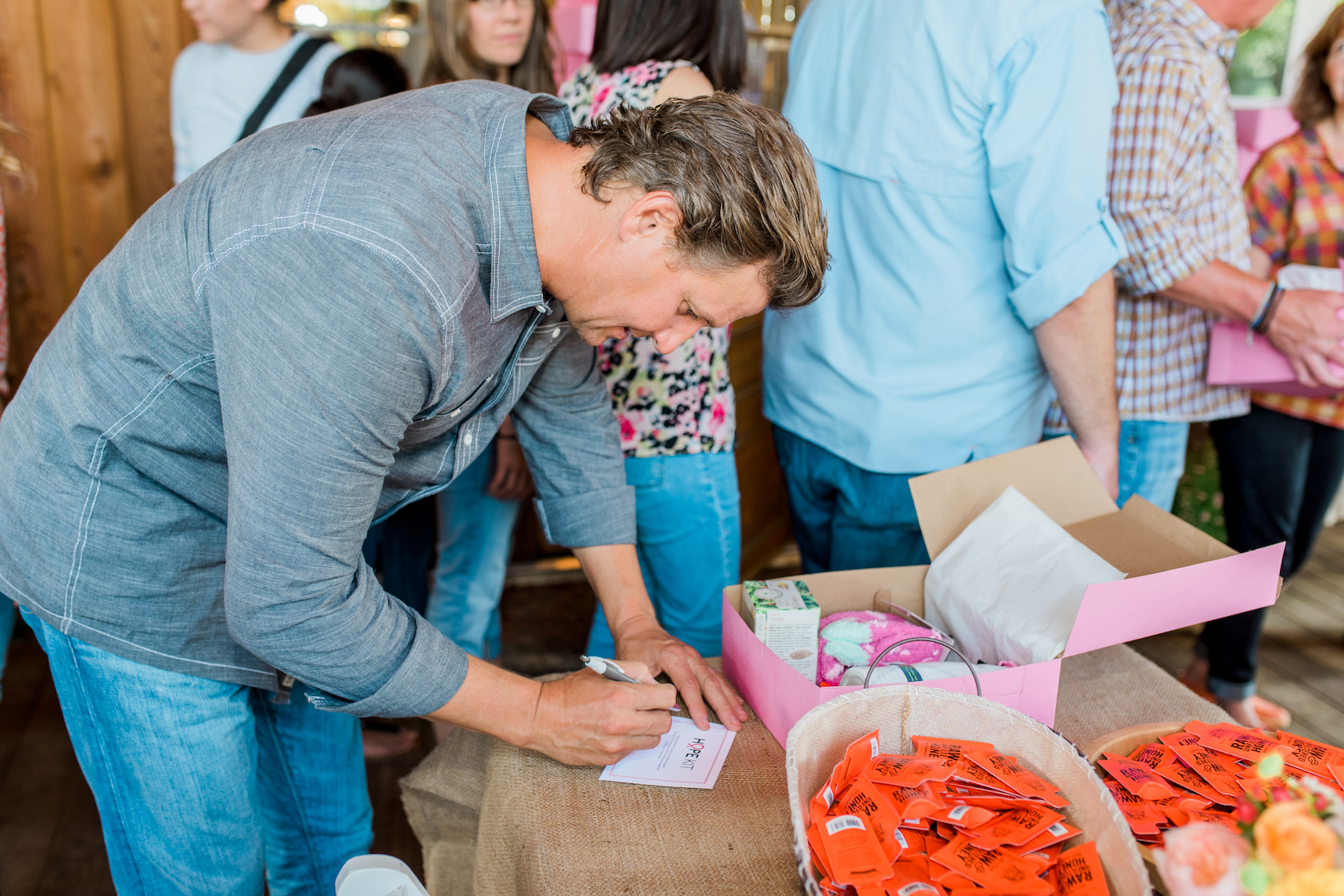 The man Nature Nate signing a card on a table with pink boxes and Nate's honey minis, with people in the background.