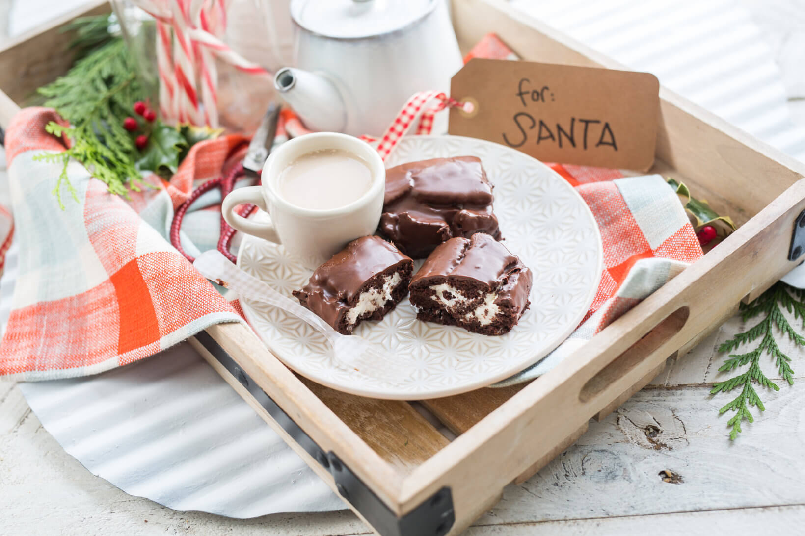 A tray with a cup of milk and chocolate-covered treats on a plate, a "for SANTA" note, surrounded by festive decoration.