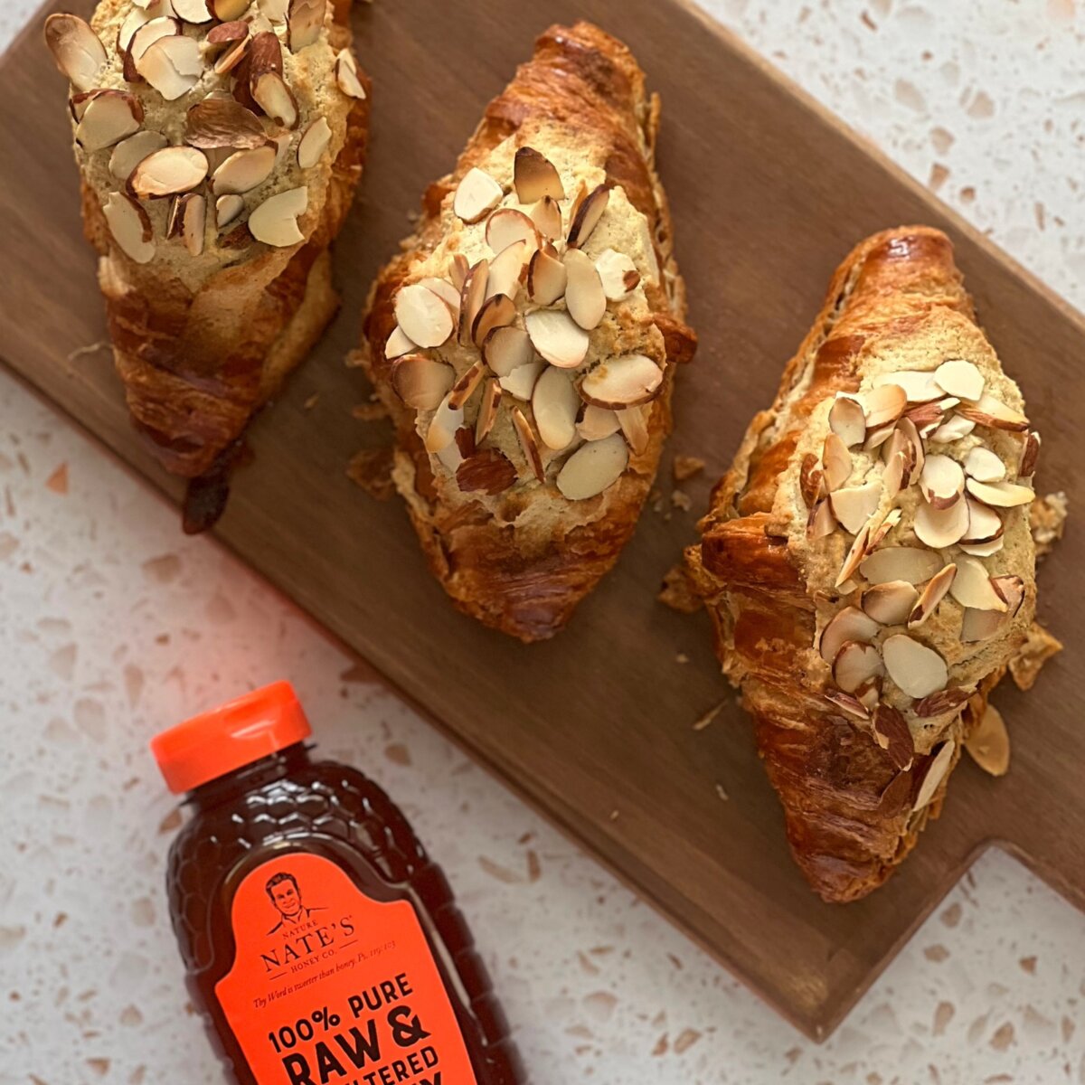 Three honey almond croissants on a board next to a Nate’s honey bottle.