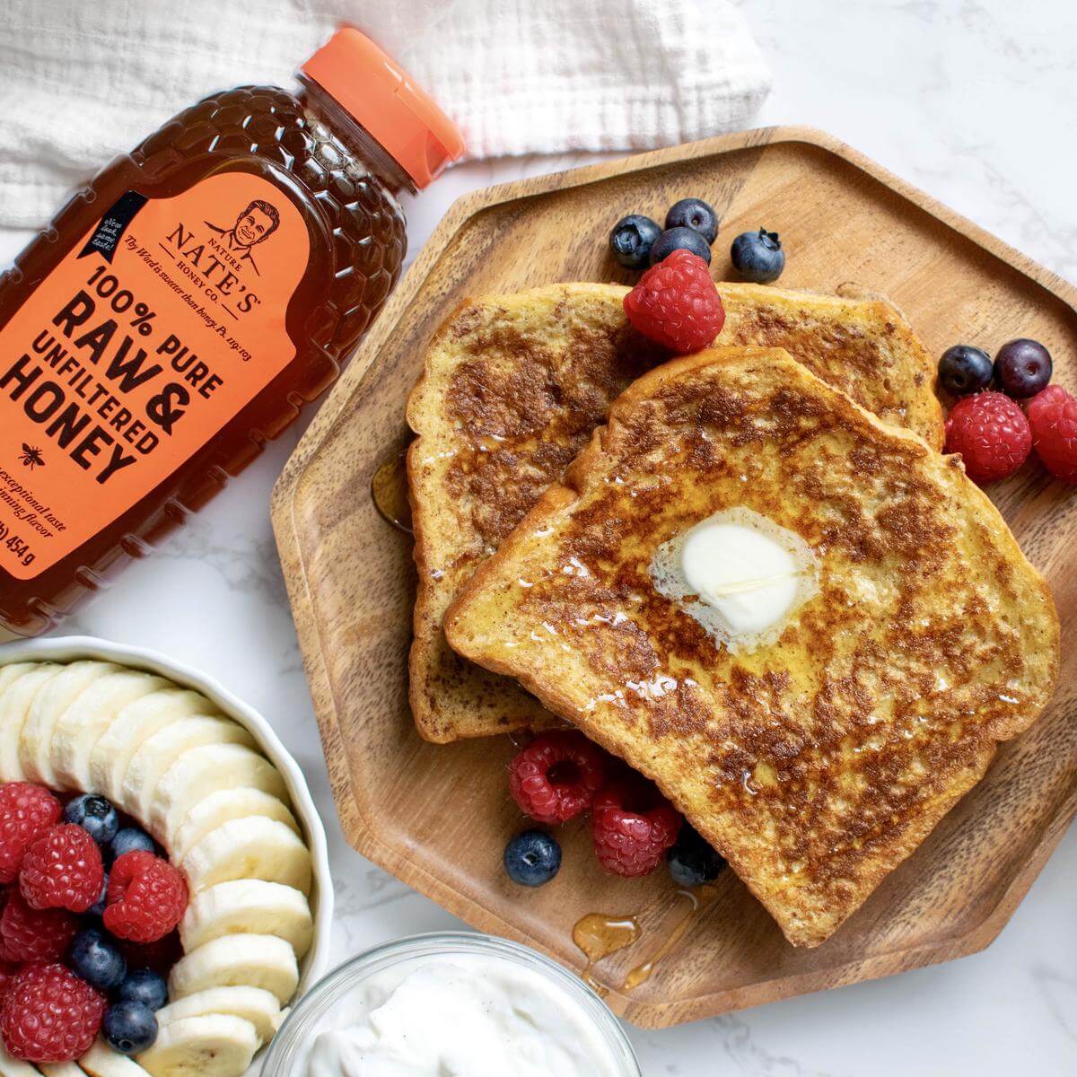 French toast with berries and banana on a plate, Nate’s honey bottle with an orange cap, on a marble background.