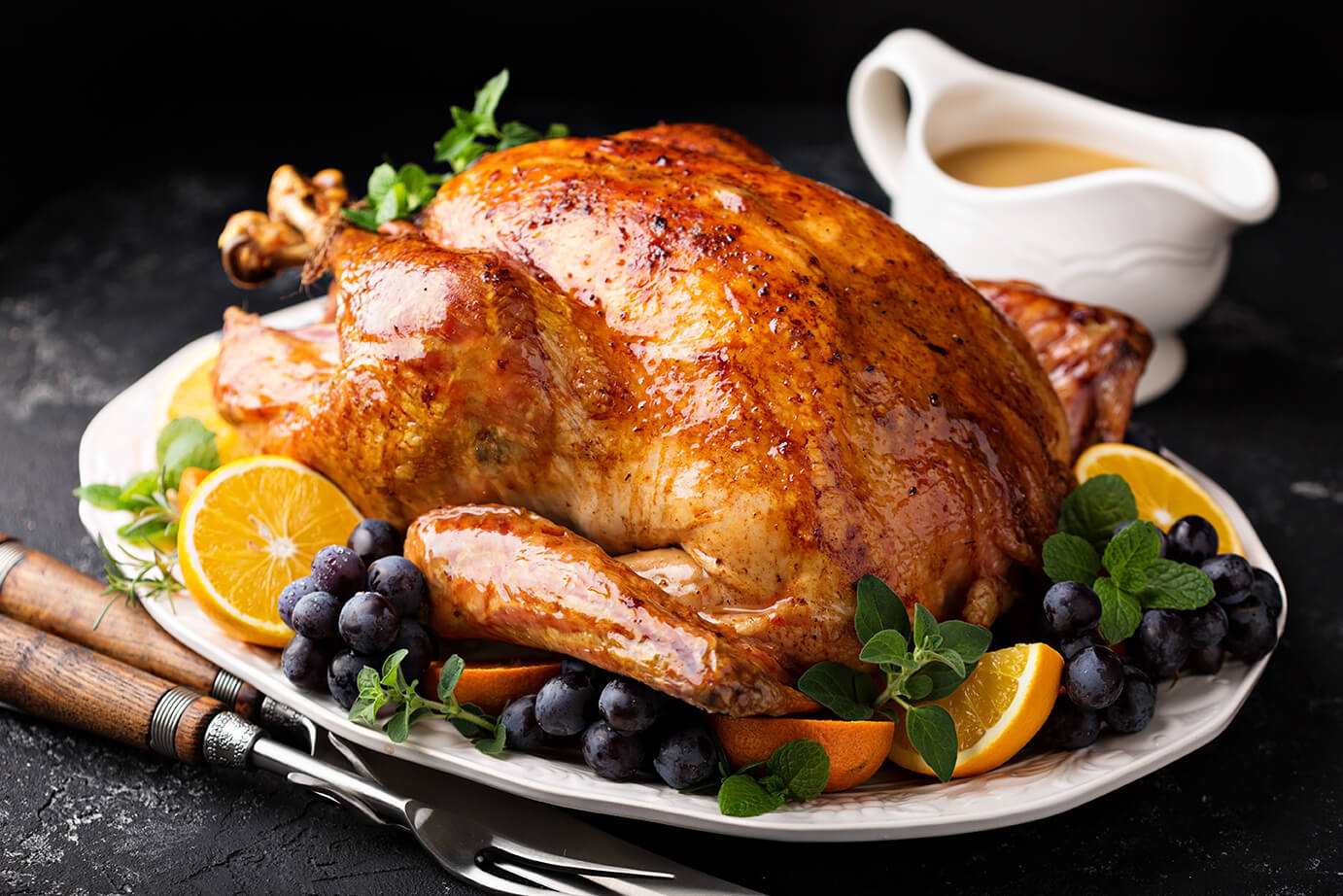 A succulent roasted turkey on a platter, garnished with orange slices, fresh grapes, and herbs, on a dark background.