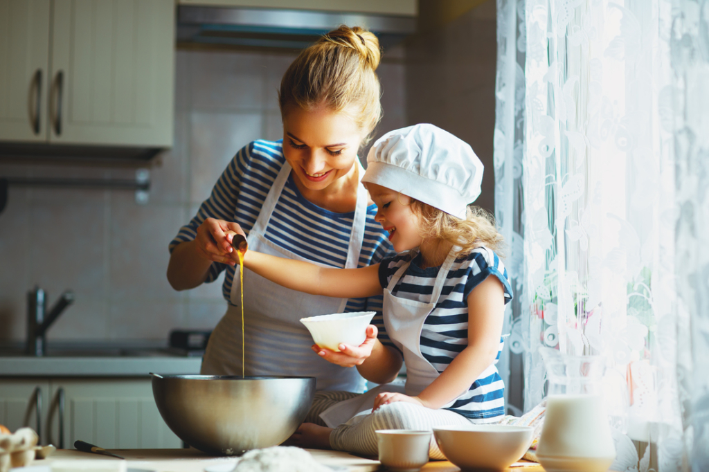 A smiling adult and child in white aprons baking together in a sunlit kitchen. The child is wearing a chef's hat.