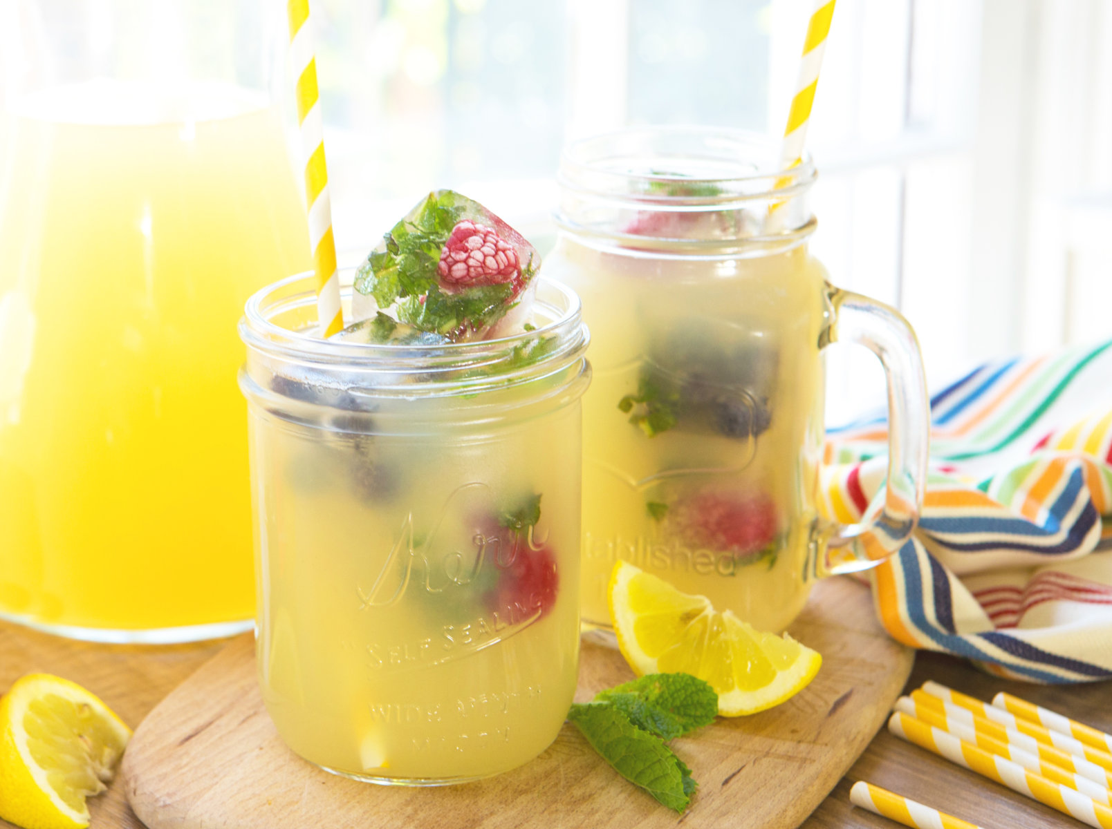 Two mason jar glasses filled with refreshing lemonade, garnished with berries and mint on a sunny table.