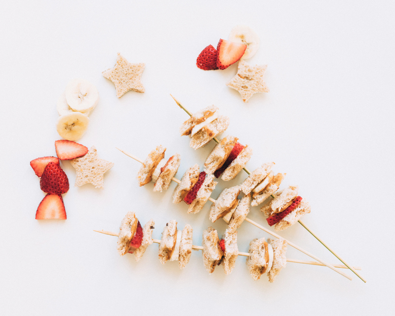 Creative food display featuring skewers of sandwich bites with various fruits arranged around in star and heart shapes.