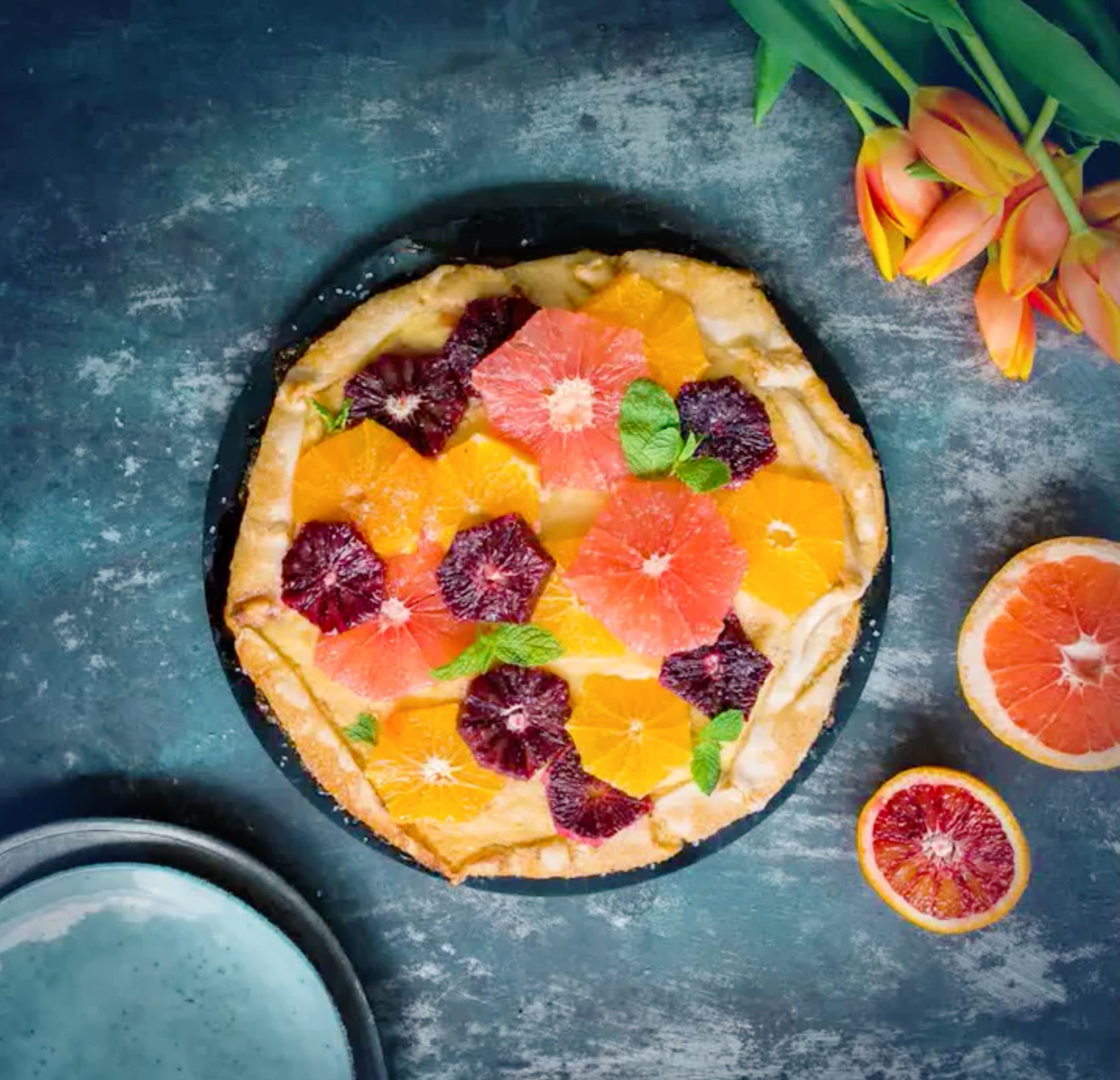 Rustic citrus tart with grapefruit slices, orange slices and mint on a blue surface, with tulips and a plate nearby.