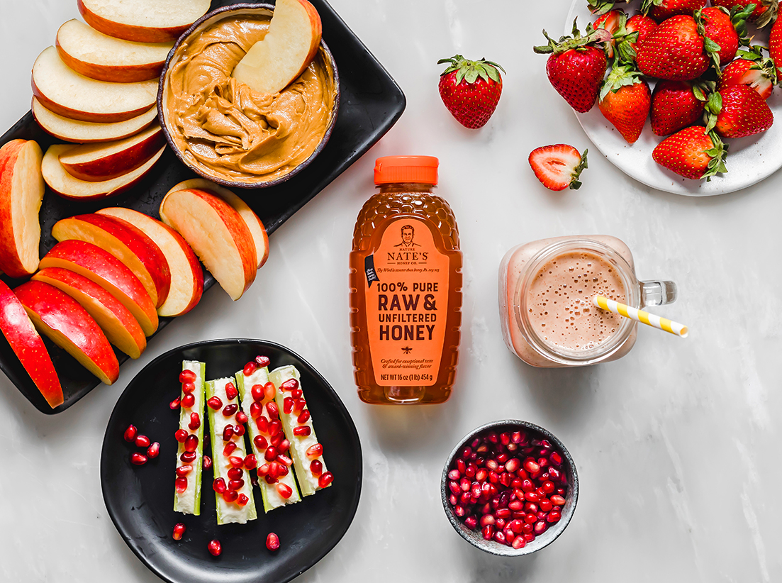 Sliced apples with peanut butter, strawberries, pomegranate seeds, a smoothie and a bottle of Nate's honey.