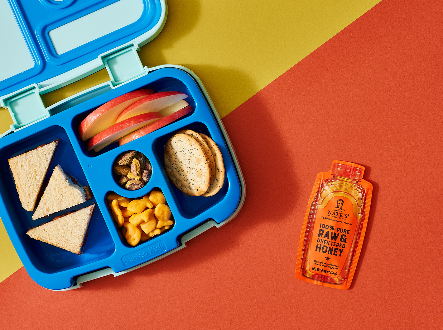 Blue bento style lunch box with apple slices, goldfish, crackers and peanut butter sandwich. Next to Nate’s honey mini.
