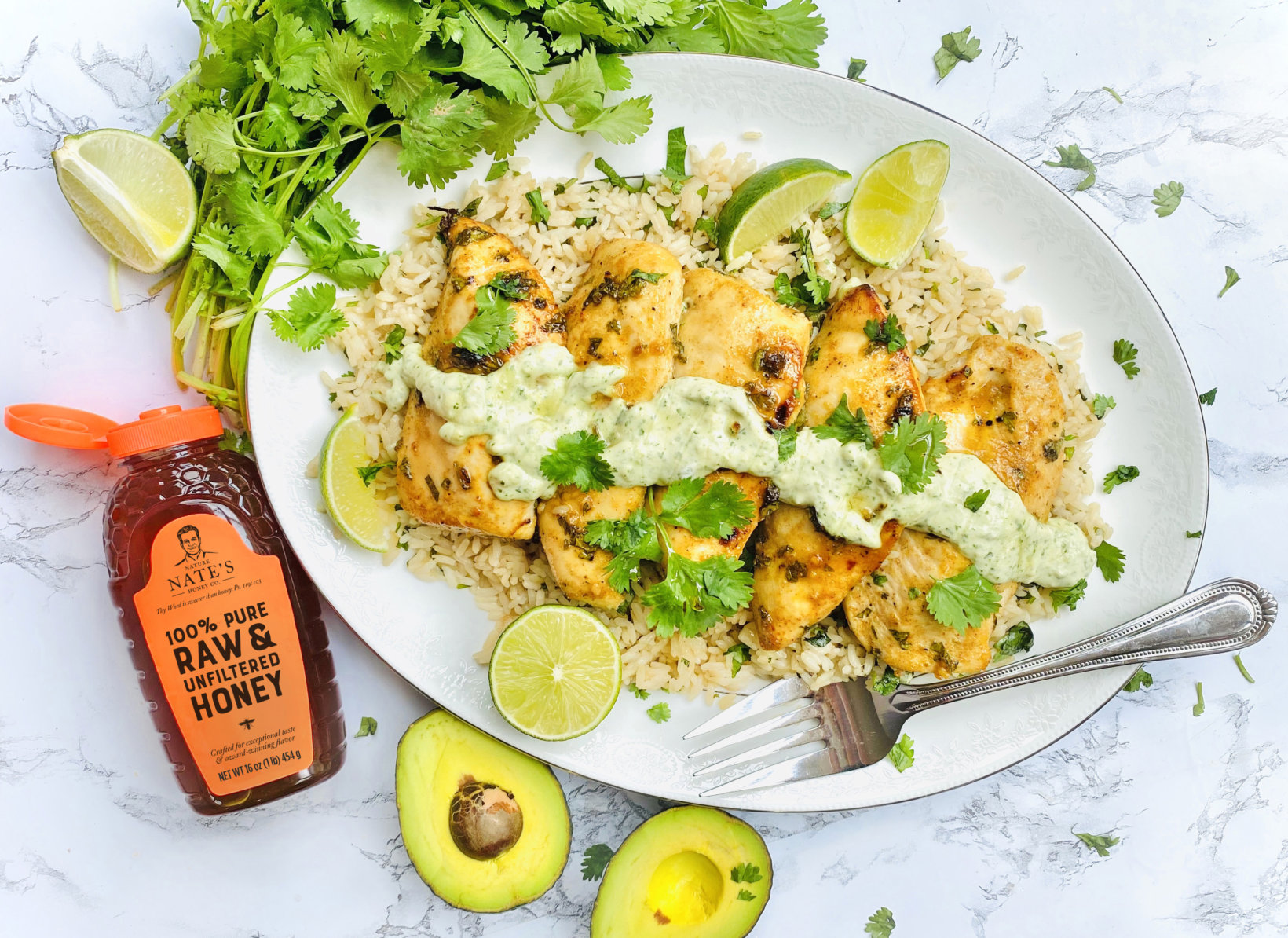 Grilled chicken on rice with honey chili sauce, avocado, lime cilantro on a platter  with a bottle of Nate’s honey.