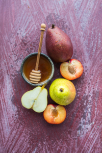 Honey with dipper in a bowl and assorted fresh fruit on a textured crimson background.