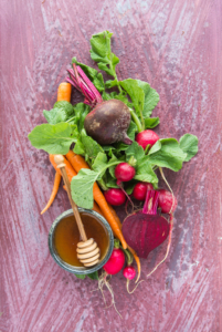 Fresh garden vegetables with honey with dipper in a bowl on a rustic pink surface.