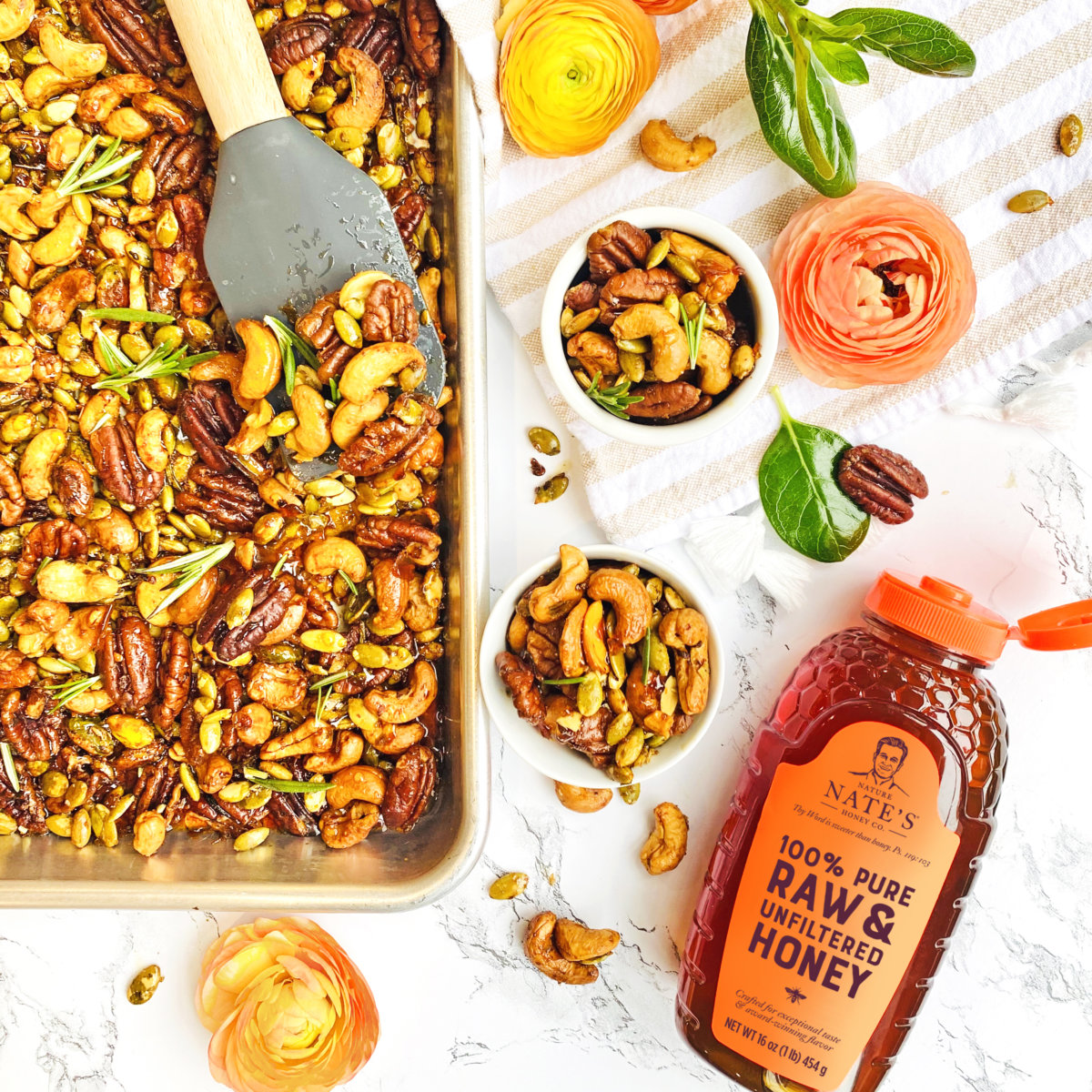 Honey-roasted nuts and seeds on a tray and in bowls with flowers and a bottle of Nate’s honey, on a marble background.