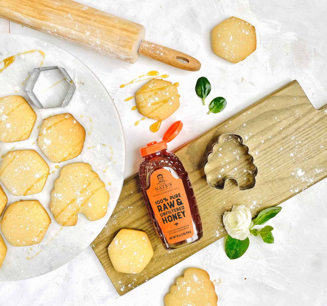 A bottle of Nate's honey with honey-drizzled cookies, rolling pin and cutters on a floured board with leaves.