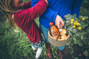 Child hugging adult holding a basket with Nate’s honey bottle, flowers and apples.