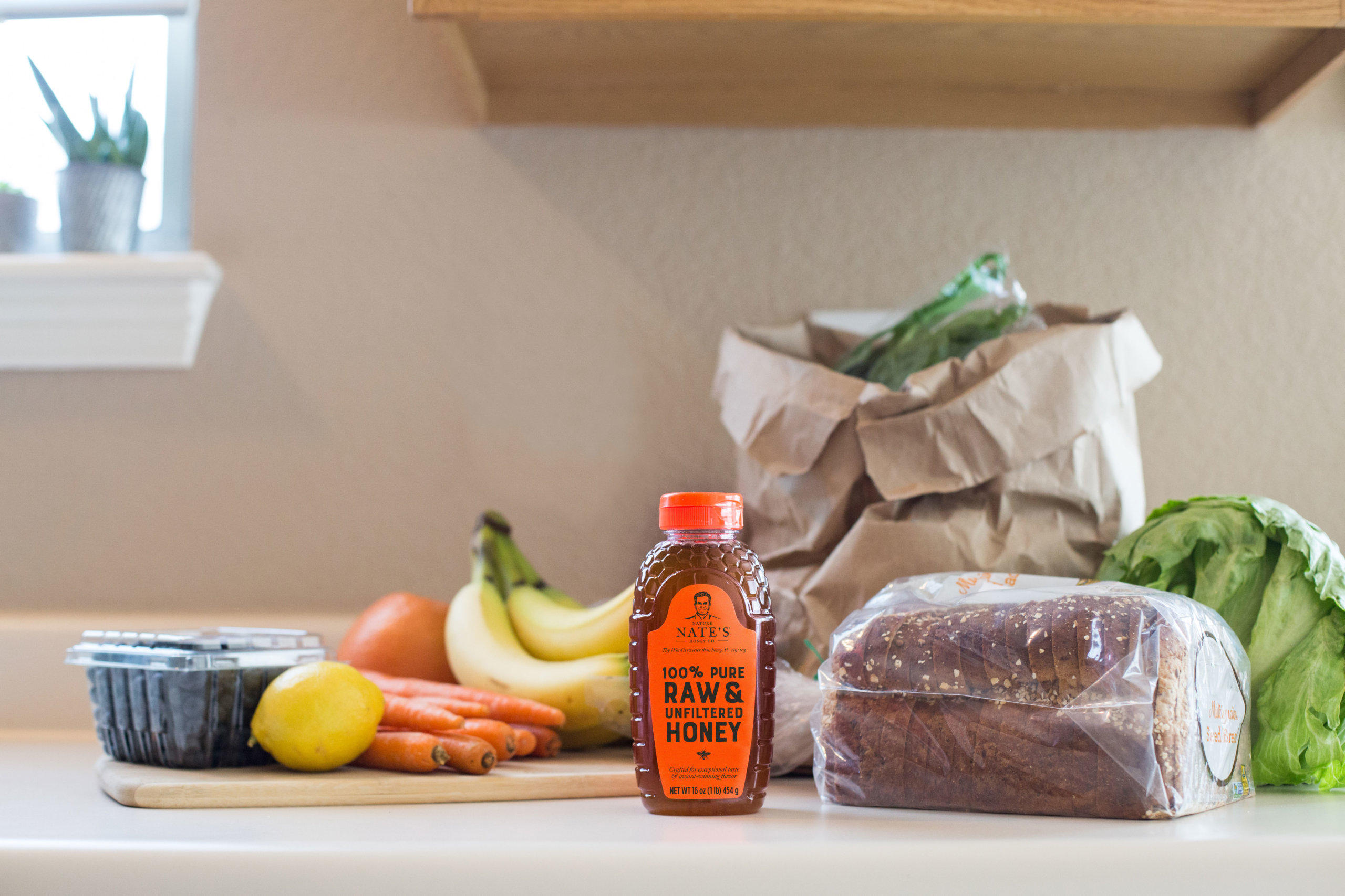 Kitchen counter with groceries including Nate's honey, fruits, carrots, lettuce and bread.