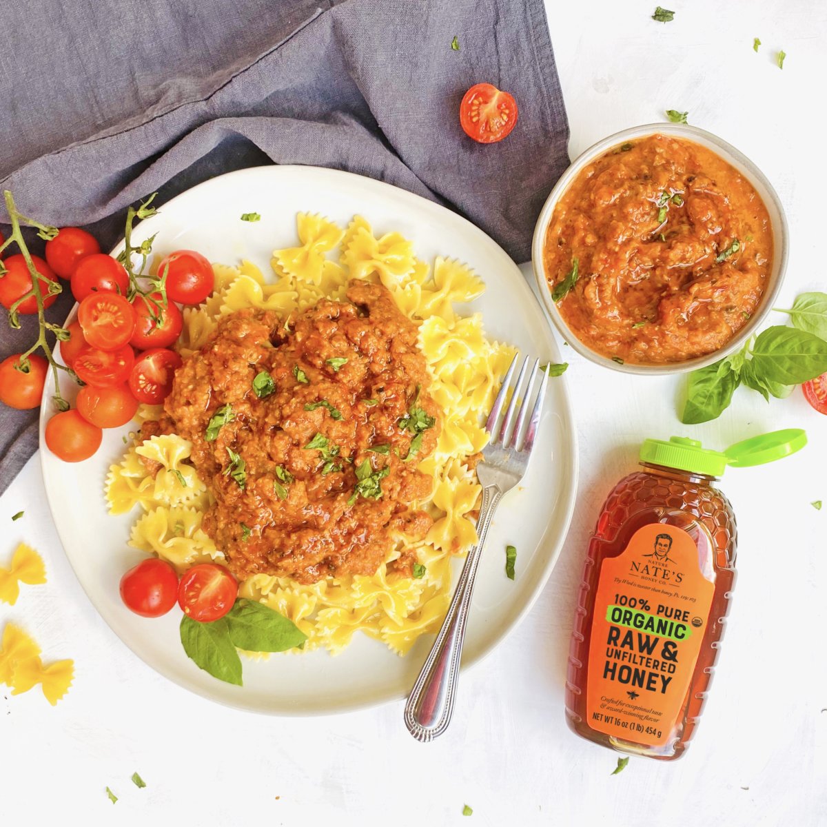 A plate of pasta with bolognese sauce, garnished with herbs, beside Nate's honey, a sauce bowl, and fresh tomatoes.