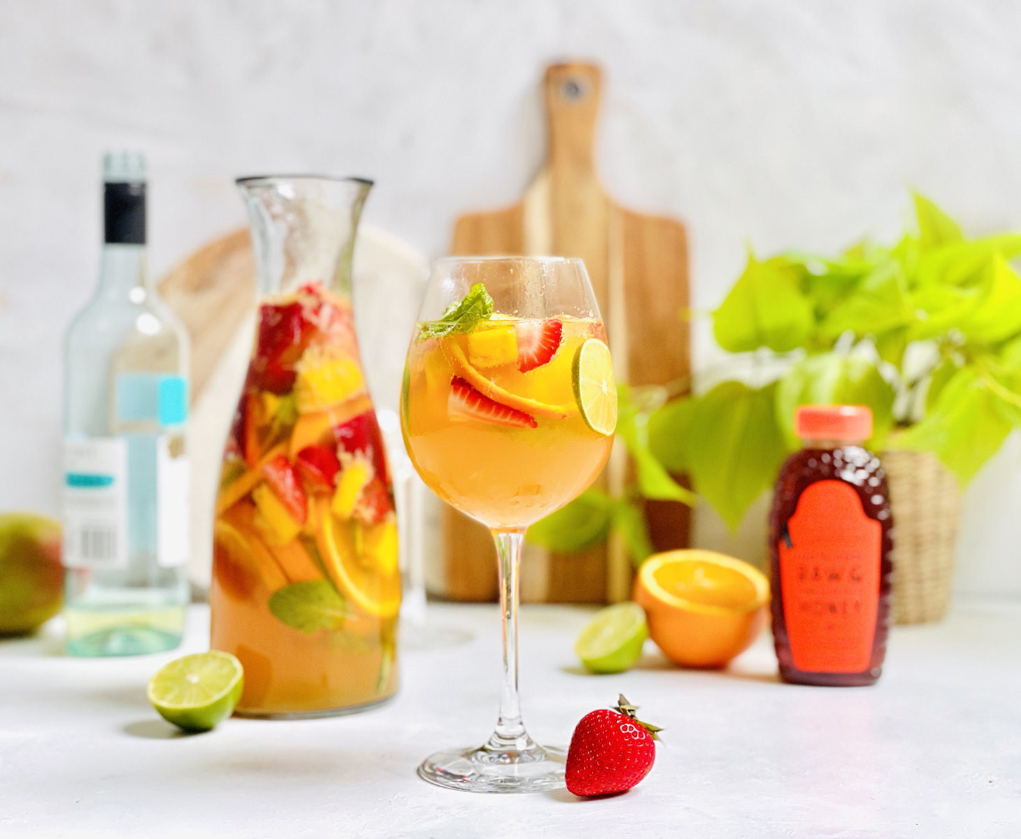 Fruity sangria in a glass and in a pitcher, spirits, a Nate’s honey bottle and fresh fruits in the background.