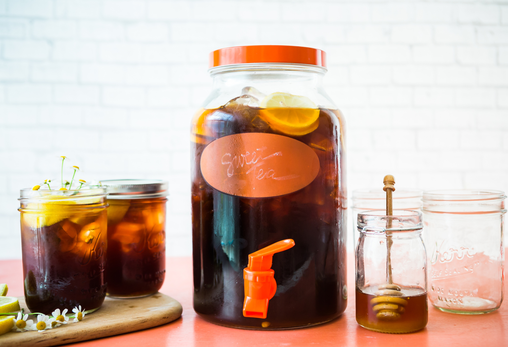 Large jar of iced tea with lemon, smaller jars, and honey with dipper on a table against a brick wall.