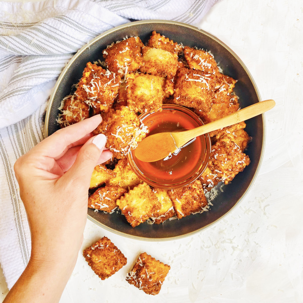 Hand dipping a honey-drizzled ravioli into a bowl of truffle honey, against a white background.