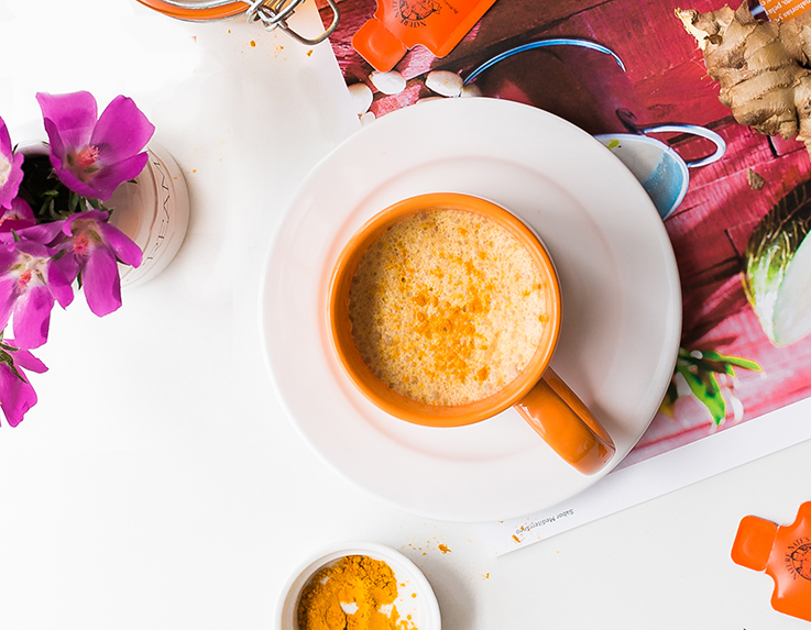 A vibrant turmeric latte in an orange cup, on a white saucer with fresh ginger and blooms and Nate's honey minis nearby.