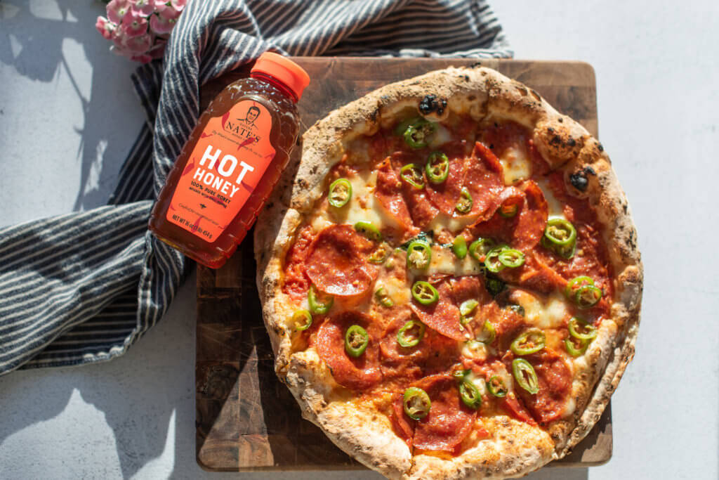 Pepperoni and jalapeño pizza on a wooden board with a bottle of Nate’s hot honey and a striped napkin beside it.