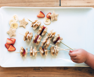 Child's hand picks skewer with banana, strawberry, sandwich bites, and star-shaped bread on white platter. Child's hand picks skewer with banana, strawberry, sandwich bites, and star-shaped bread on white platter.
