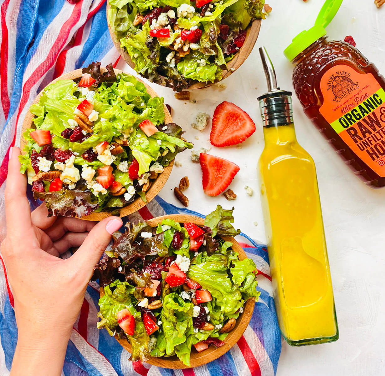 Hand holding a berry and feta salad, with a bottle of dressing and a bottle of Nate’s organic honey on a striped towel.
