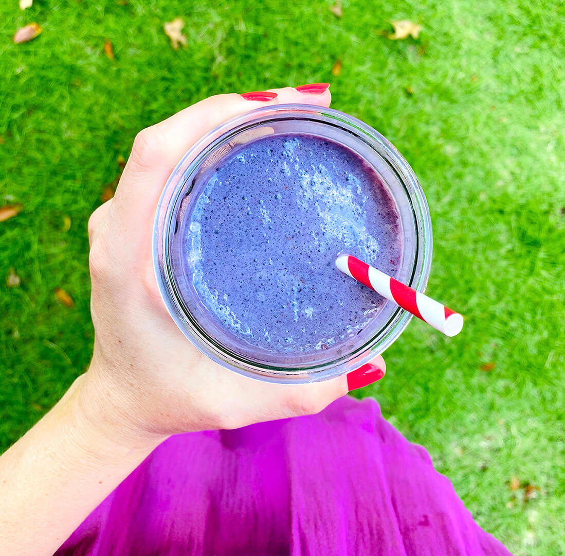 Hand holding a blue smoothie with a straw, over a green lawn and purple dress.