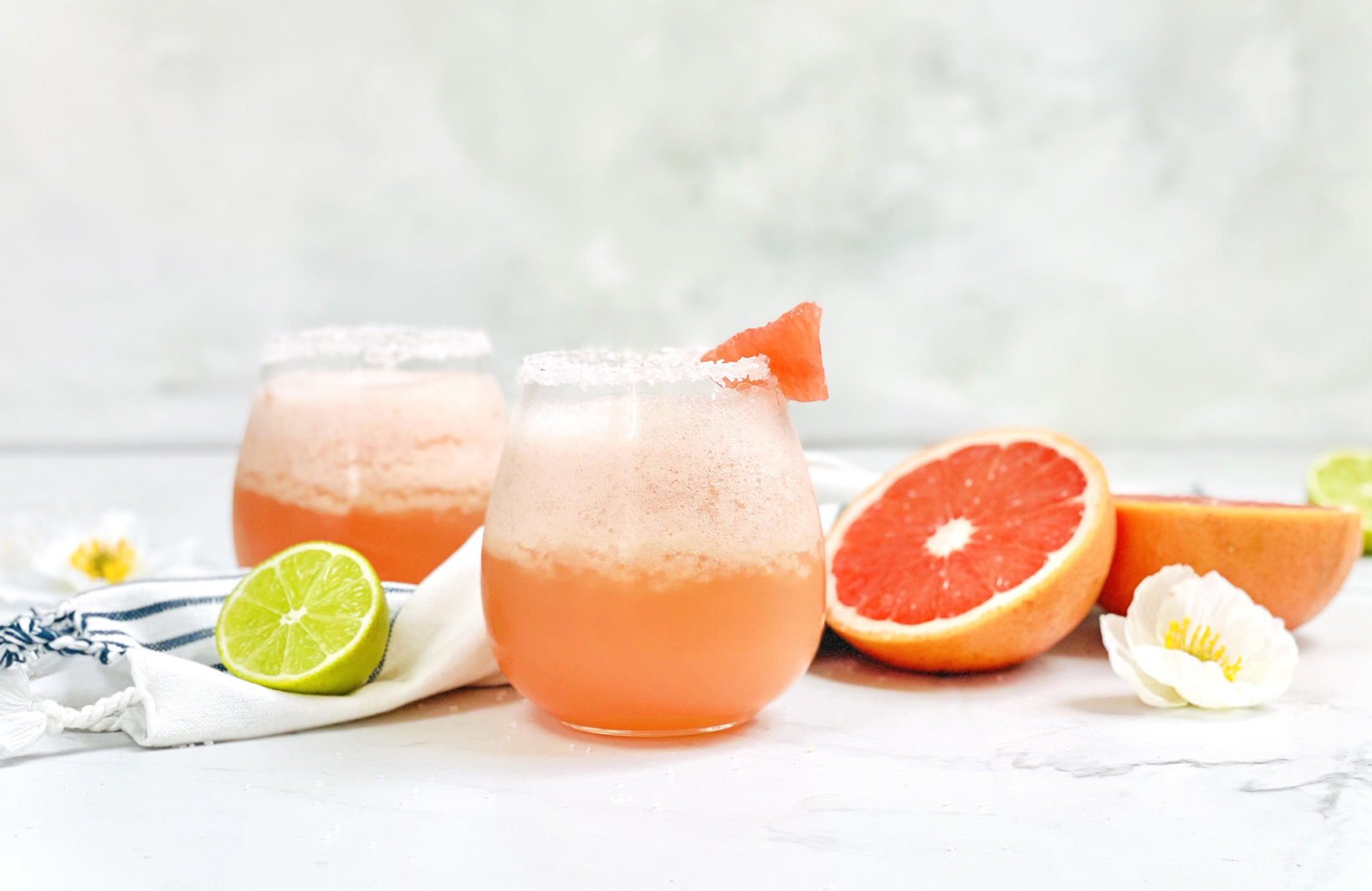 Refreshing frozen paloma cocktails, served on a light surface with fresh citrus fruits and flowers nearby.