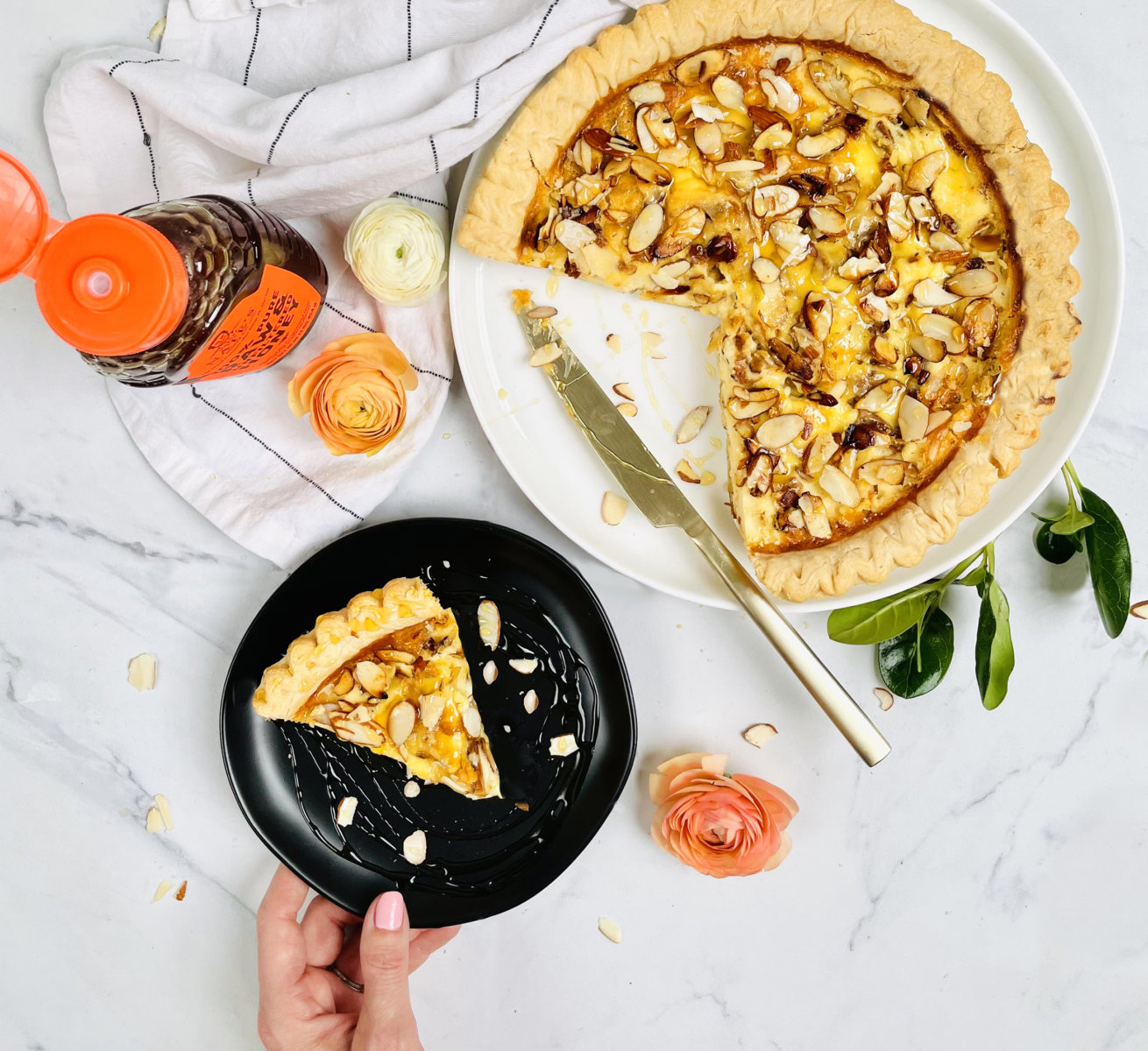 Honey Almond Sweet Quiche on platter and on black plate next to bottle of Nate's honey, flowers and white towel on marble background