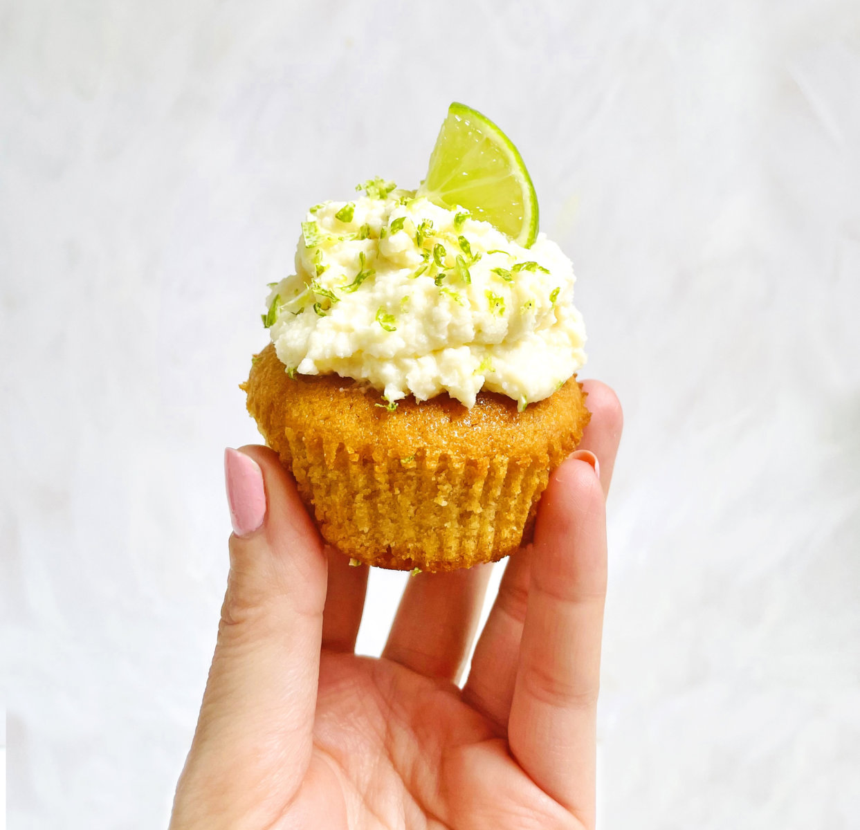 A hand holding a cupcake topped with cream and lime zest against a white background.