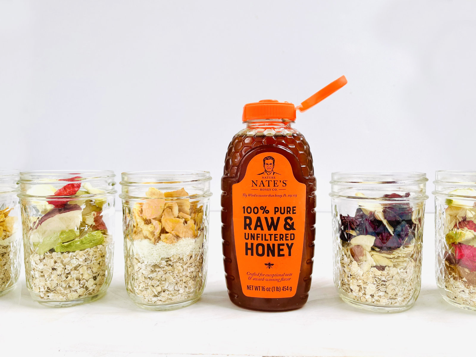 Nate's honey bottle with glass jars filled with oatmeal and assorted fruit on a white backdrop.