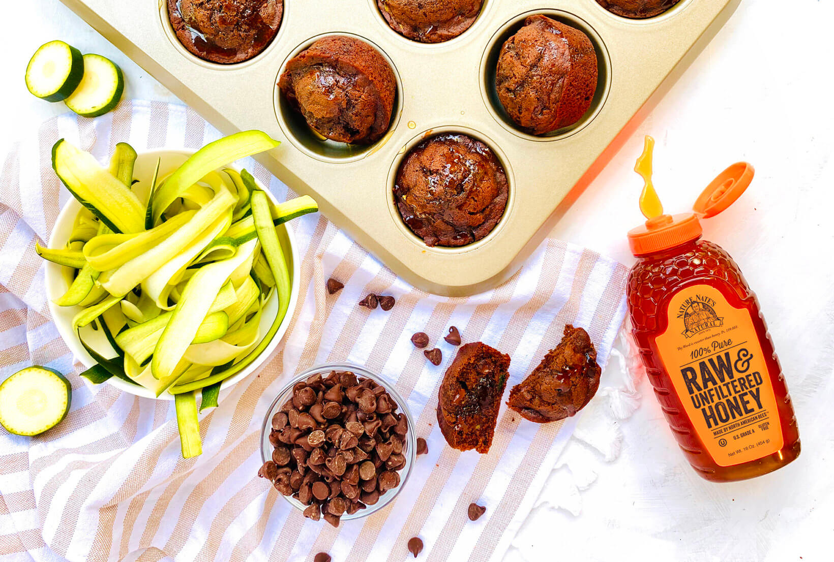 Chocolate muffins in a pan, Nate’s honey bottle, chocolate chips and zucchini ribbons on a striped towel.