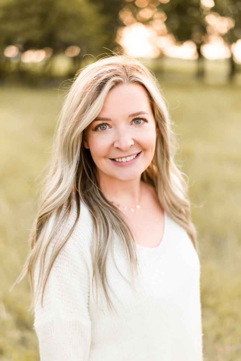 Smiling woman in white sweater with long blonde hair standing in field with trees in background.