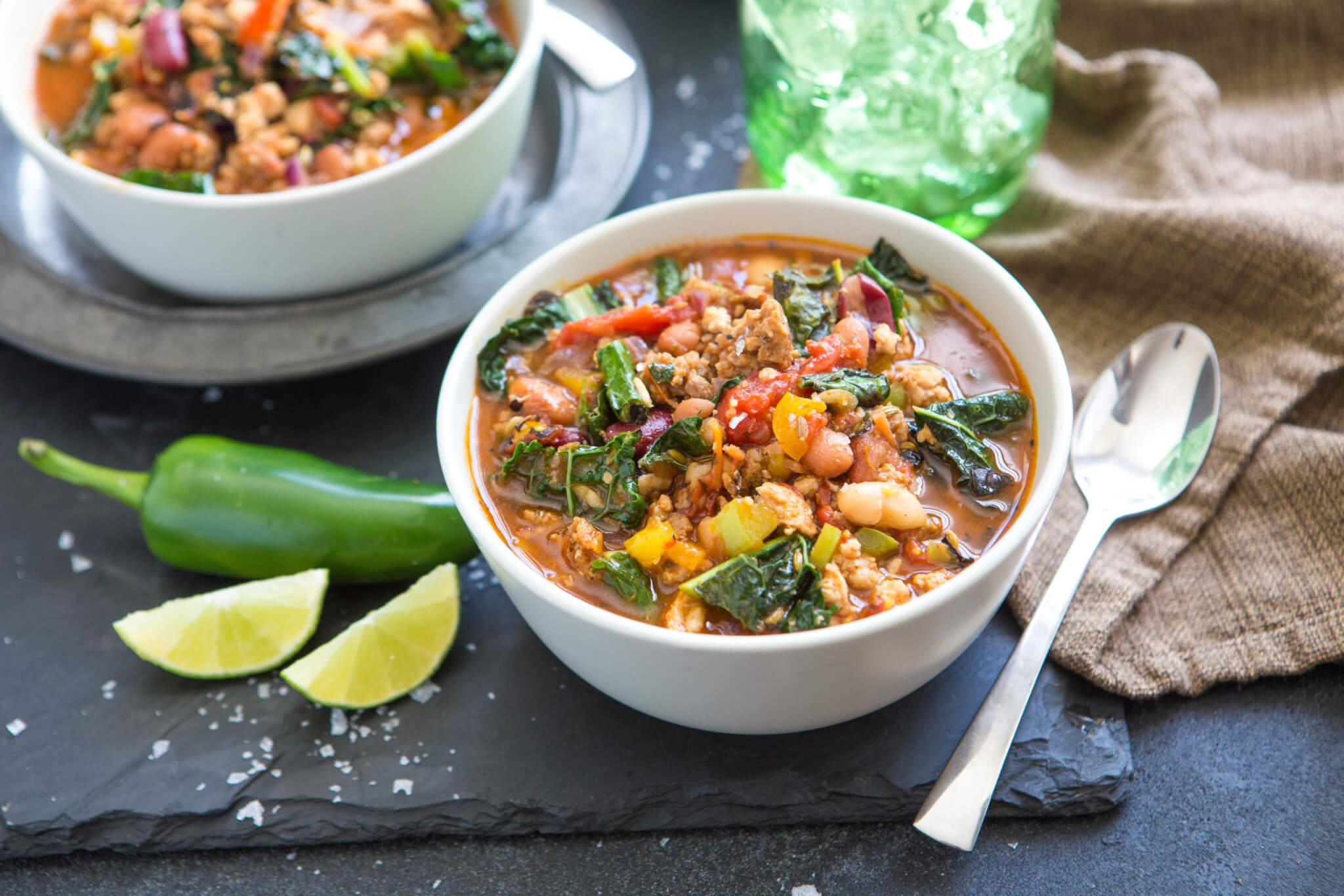 A bowl of hearty stew with greens, spoon and lime on slate.